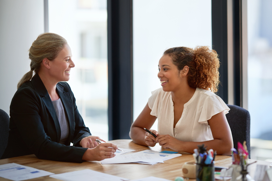 Two people talk at a desk in an office. Papers and pens are on the desk.