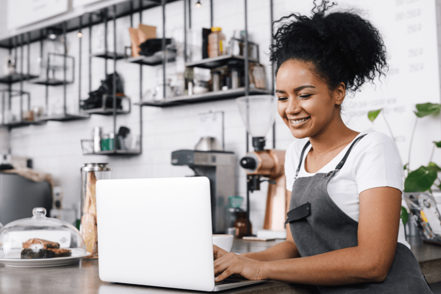 A women sits behind the counter of a coffee shop and looks at information on a laptop.